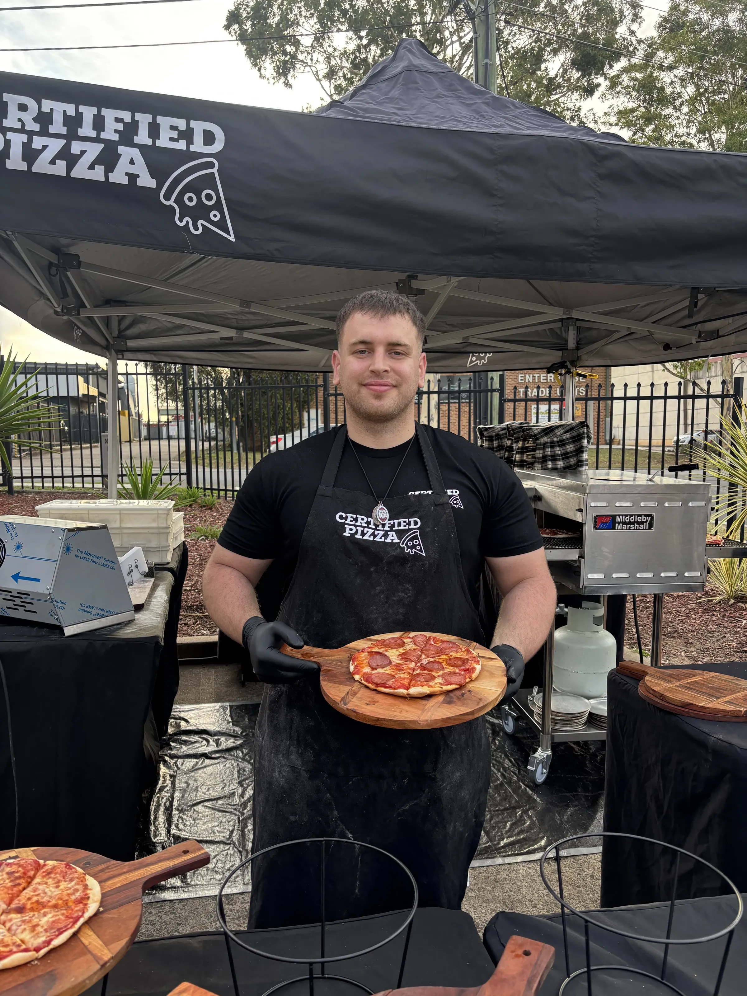 Certified Pizza team member holding a fresh pepperoni pizza beside the commercial conveyor oven in Narellan